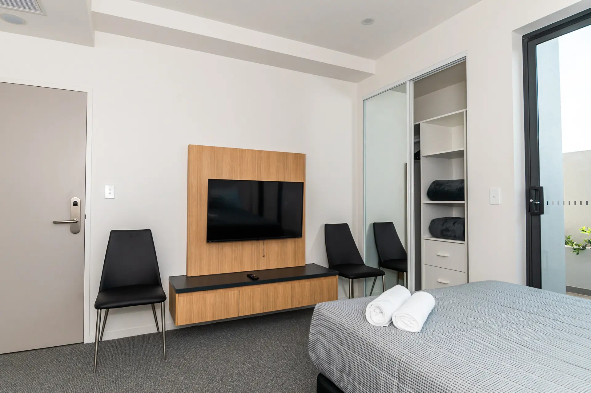 Apartment bedroom with TV on timber feature wall, mirrored wardrobe and balcony access
